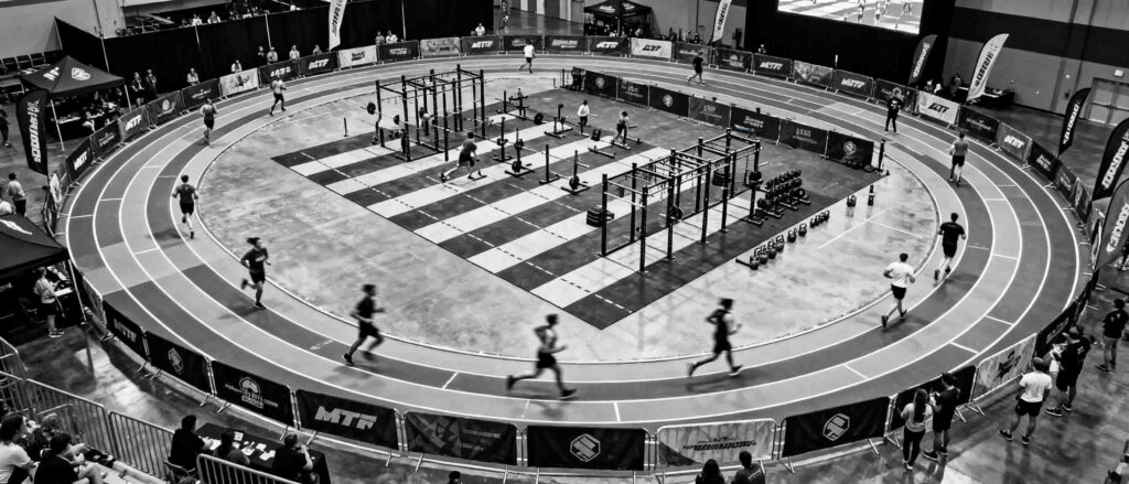 Black‑and‑white aerial view of an indoor HYROX arena, showing athletes running on an oval track surrounding a central workout zone filled with rigs, weights, and functional training stations, with spectators around the edges.