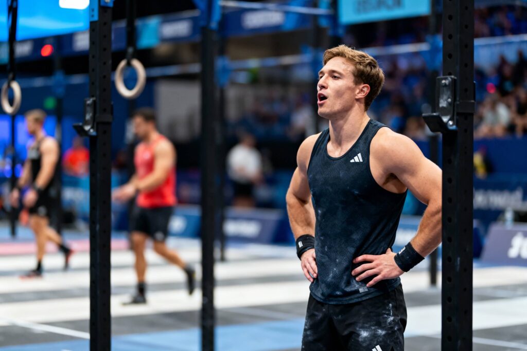 Color photo of an athlete standing with hands on hips between pull-up rig posts in a competition arena, chalk on his dark tank top and other competitors blurred in the background.
