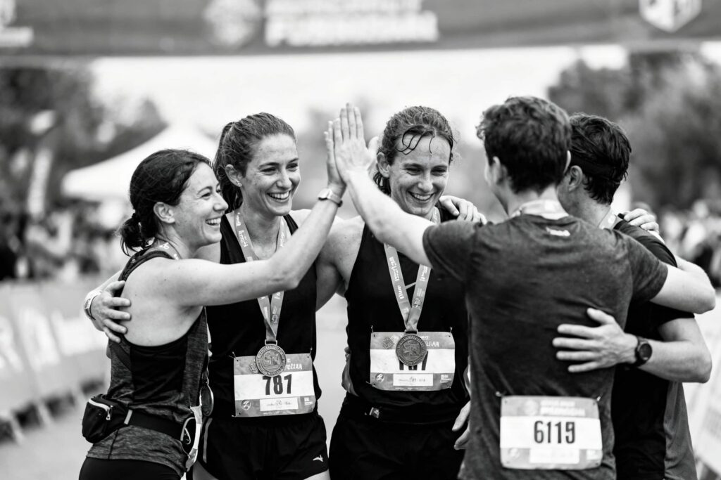 Black‑and‑white photo of a group of runners wearing race bibs and medals, standing together at the finish line and celebrating with high‑fives and hugs.
