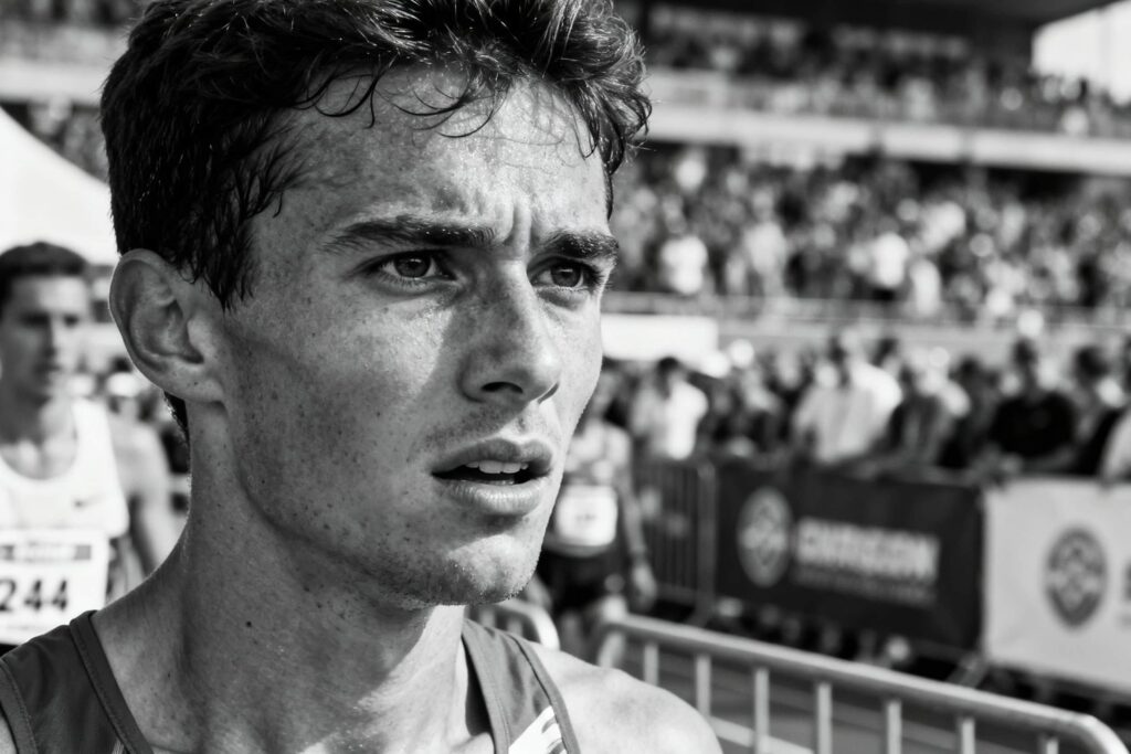 Black‑and‑white close‑up of a sweaty runner at a race finish area, with a blurred crowd and other competitors behind the barrier in the background.