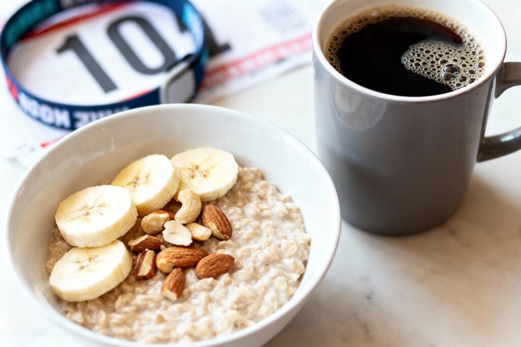 Close‑up of a pre‑race breakfast showing a bowl of oatmeal topped with banana slices and mixed nuts next to a mug of black coffee, with a race bib and lanyard blurred in the background.